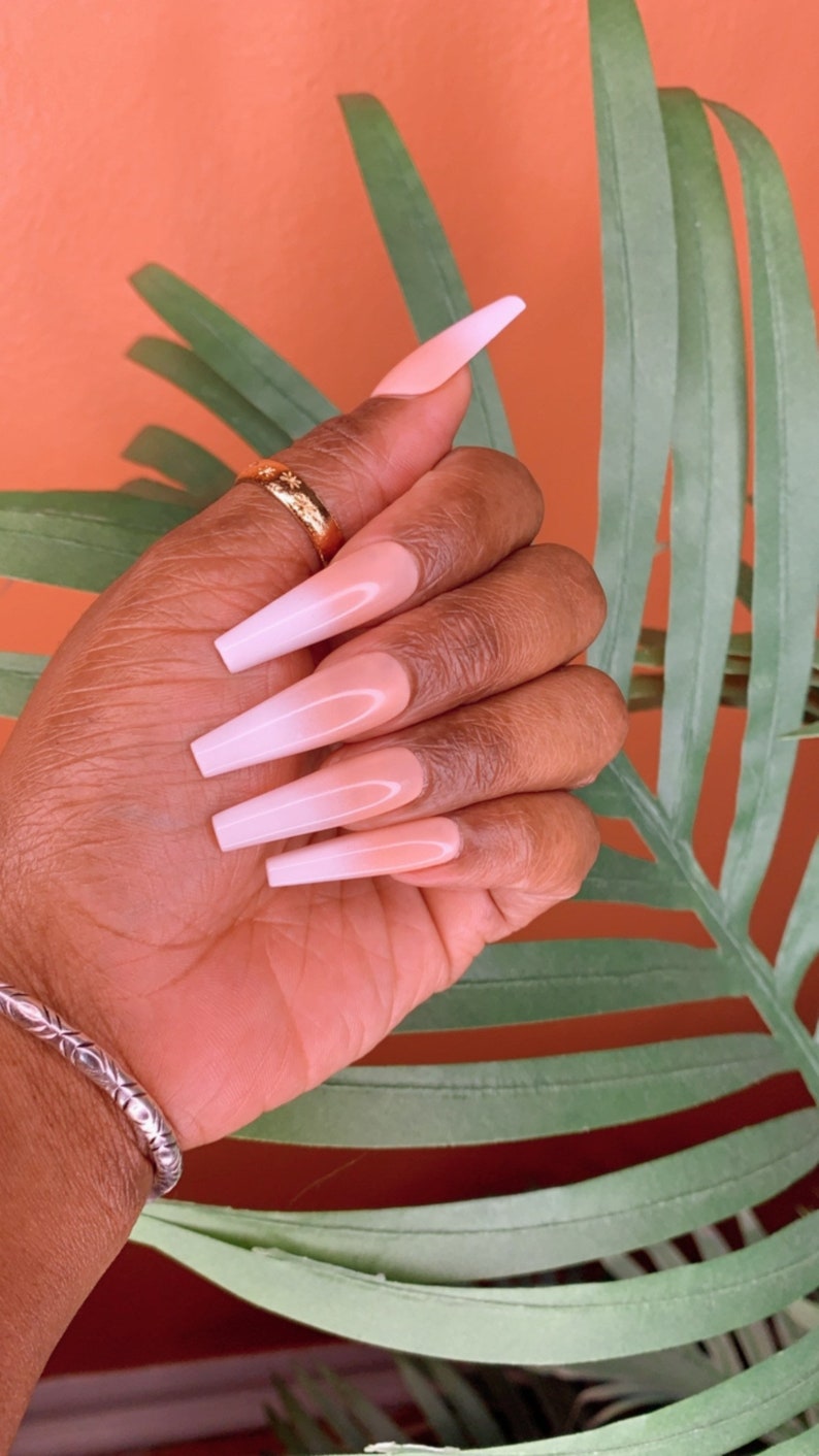 May include: Close-up of a hand with long, almond-shaped nails. The nails are painted with a white ombre design, transitioning from a light pink base to a white tip.