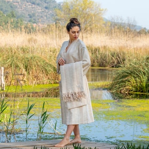 May include: A woman wearing a white robe with a striped pattern stands on a wooden dock over a pond. She is holding a white towel with fringe.