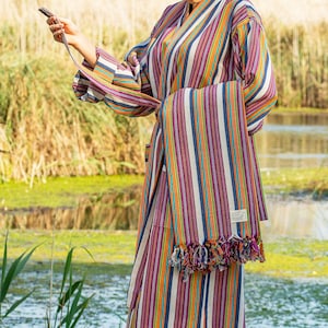 May include: A woman wearing a colorful striped robe with a matching striped towel draped over her arm. The robe has a sash tie and the towel has fringe. The woman is standing on a wooden dock by a body of water.
