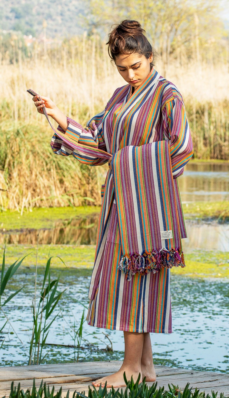 May include: A woman wearing a colorful striped robe with a matching striped towel draped over her arm. The robe has a sash tie and the towel has fringe. The woman is standing on a wooden dock by a body of water.