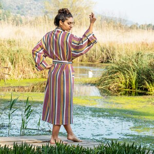 May include: A woman wearing a long, striped robe with a sash. The robe is in shades of pink, yellow, blue, and white. She is standing on a wooden dock over a pond with green reeds and water.