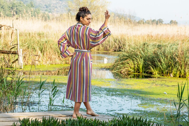 May include: A woman wearing a long, striped robe with a sash. The robe is in shades of pink, yellow, blue, and white. She is standing on a wooden dock over a pond with green reeds and water.