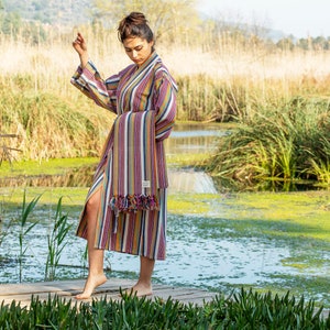 May include: A woman wearing a long, striped robe with a fringe trim. The robe is in shades of pink, yellow, blue, and white. She is standing on a wooden dock over a pond with green reeds in the background.