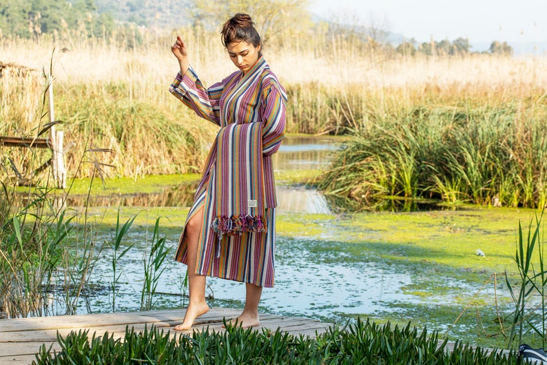 May include: A woman wearing a long, striped robe with a fringe trim. The robe is in shades of pink, yellow, blue, and white. She is standing on a wooden dock over a pond with green reeds in the background.