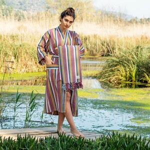 May include: A woman wearing a striped robe holds a matching striped towel in front of a pond with green algae and tall grass.