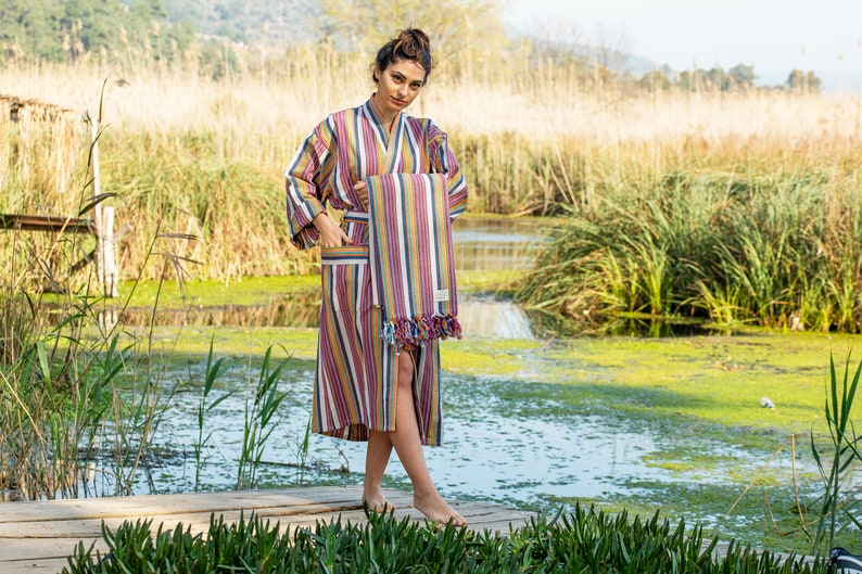 May include: A woman wearing a striped robe holds a matching striped towel in front of a pond with green algae and tall grass.