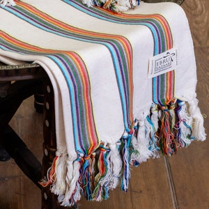 Puede incluir: Toalla turca blanca con rayas de colores y flecos. La toalla está doblada y drapeada sobre una silla de madera. La toalla tiene una etiqueta que dice "Ebru Bazaar".
