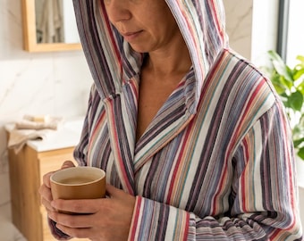 May include: A hooded bathrobe featuring a vertical striped pattern in shades of red, blue, white, and grey. The robe includes long sleeves, a tie waist, and two front pockets. A solid tan mug is held in the hands, completing the loungewear look.