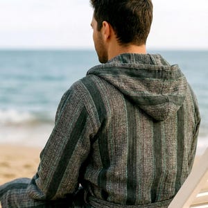 May include: A hooded robe with vertical stripes in shades of gray and green. The robe has a belt and is worn by a person sitting on a beach chair. The background shows the ocean and a sandy beach.