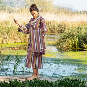 May include: A woman wearing a colorful striped robe with fringe trim stands on a wooden dock over a pond. The robe is tied at the waist and has a long, flowing design. The pond is surrounded by tall grass and reeds.