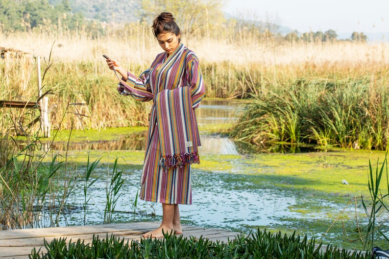 May include: A woman wearing a colorful striped robe with fringe trim stands on a wooden dock over a pond. The robe is tied at the waist and has a long, flowing design. The pond is surrounded by tall grass and reeds.