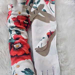 Puede incluir: Guantes blancos con un vibrante estampado floral que presenta grandes amapolas rojas, hojas verdes y pequeñas flores blancas. Los guantes son largos, se extienden hasta el antebrazo y son de tela.