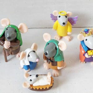 May include: A collection of ten small clay figurines depicting a nativity scene with mice. The set includes mice dressed in colourful robes, a mouse with purple wings and a halo, mice wearing crowns, and a small white sheep figurine.