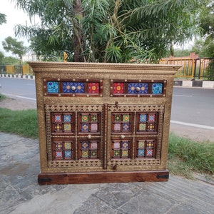 Puede incluir: Un armario de madera con dos puertas y dos cajones. El armario está decorado con intrincados incrustaciones de latón y azulejos coloridos. El armario es de un color marrón intenso y tiene un acabado desgastado.