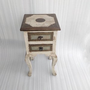 May include: White wooden nightstand with two drawers and ornate metal accents. The top drawer has a decorative design with a circular pattern in the center.