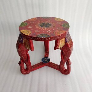 May include: A red wooden stool with three elephant legs. The top of the stool is round and decorated with colorful mandala patterns.