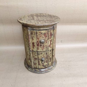 May include: A round wooden side table with two drawers. The table is painted in a distressed green and brown finish. The drawers have metal handles.