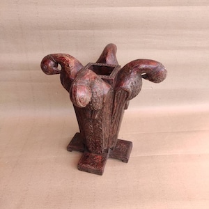 May include: A dark brown wooden sculpture of a vase with four bird heads. The vase has a square opening at the top and a square base.