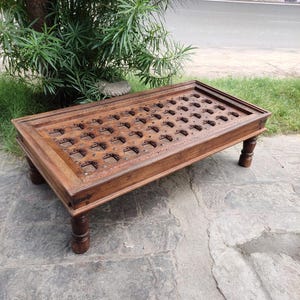 Puede incluir: Una mesa de centro de madera marrón oscuro con tallas intrincadas y una tapa rectangular. La mesa tiene cuatro patas torneadas y se encuentra en un patio de piedra.
