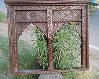 Può includere: Una cornice in legno marrone scuro con intagli intricati e due aperture ad arco. La cornice è appoggiata su una base in pietra davanti a erba verde.