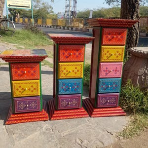 May include: Three colorful, hand-painted wooden drawer chests. Each chest features a red base and top with intricate designs, and drawers in various colors like orange, yellow, blue, pink, and purple. The chests are adorned with decorative knobs.