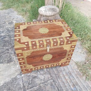 May include: A wooden chest with brass accents and intricate carvings. The chest has a hinged lid and a brass lock. The chest is sitting on a stone surface.