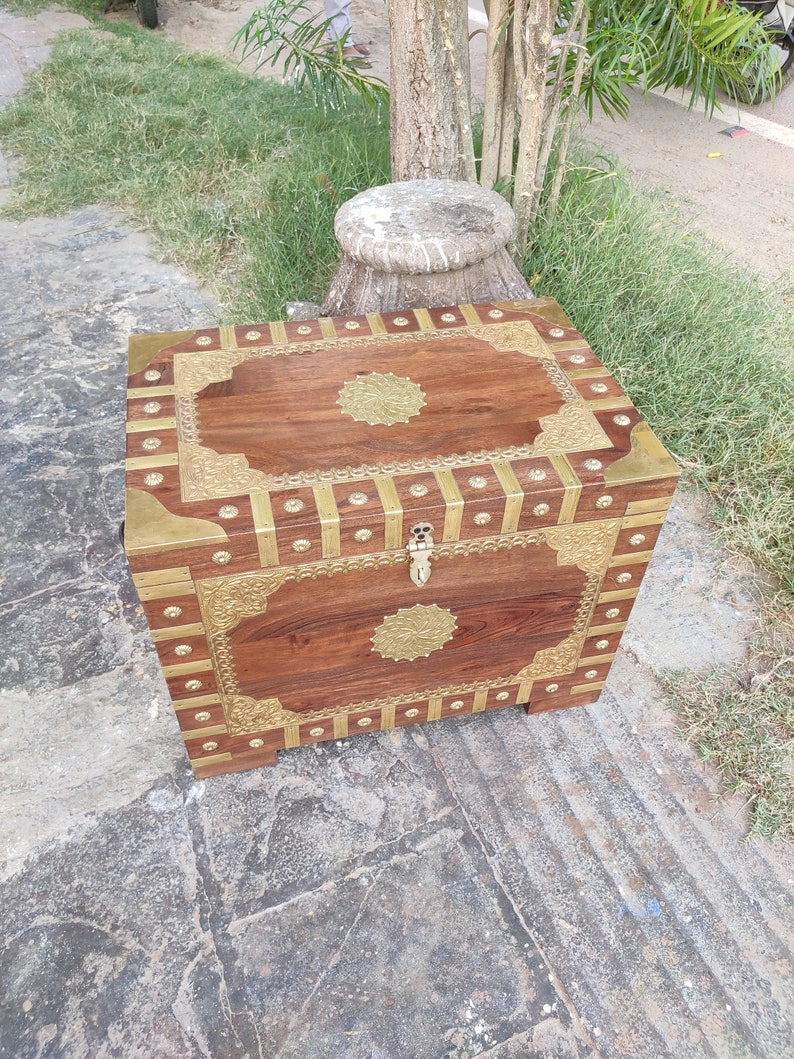 May include: A wooden chest with brass accents and intricate carvings. The chest has a hinged lid and a brass lock. The chest is sitting on a stone surface.