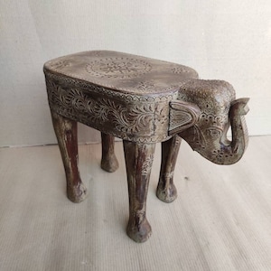 May include: A carved wooden elephant stool with intricate details. The stool has a round top and four legs shaped like elephant legs. The elephant's trunk is curled up and the eyes are carved with detail.