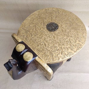 May include: A gold and brown wooden elephant stool with an ornate, engraved top. The top is round and has a small, circular design in the center.