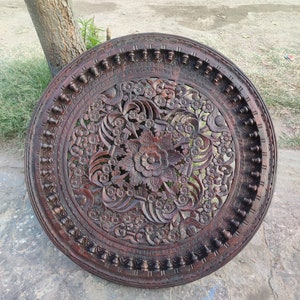 May include: A round, dark brown wooden wall decor with intricate carvings of flowers and leaves. The design is symmetrical and features a central floral motif.