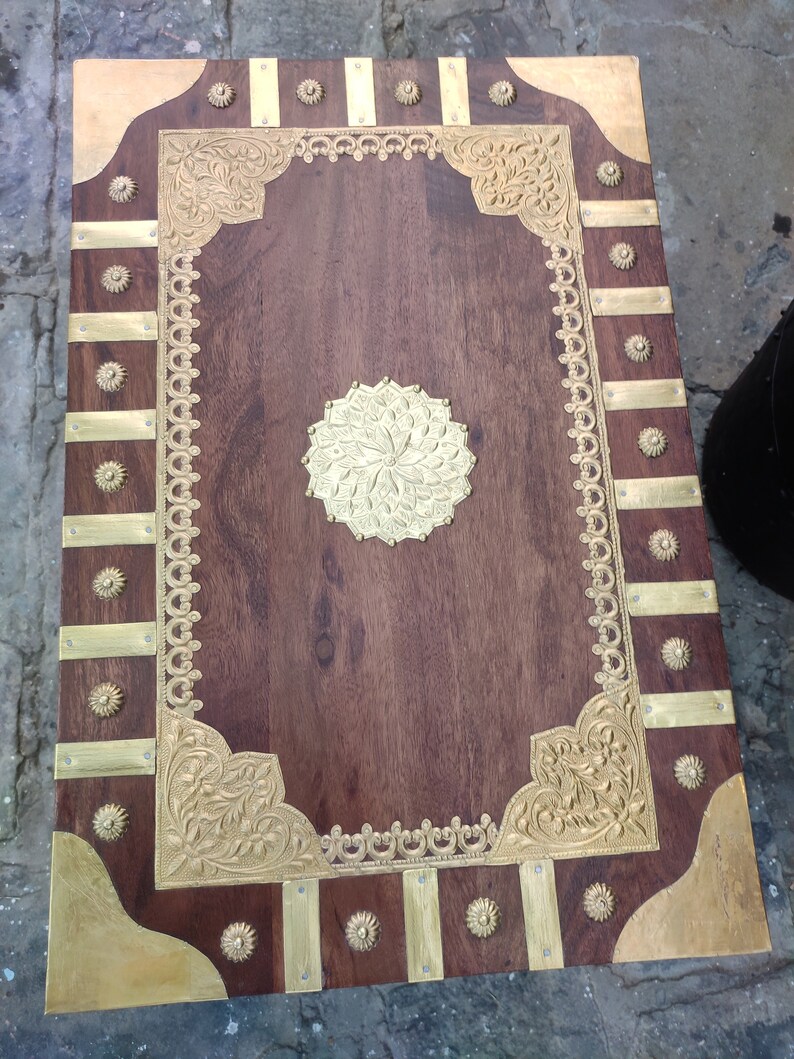May include: A wooden box with a decorative brass inlay. The box has a dark brown wood finish and a brass border with intricate floral designs. The top of the box features a large brass circle with a floral design.