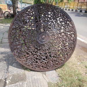 May include: A large, round, dark brown wooden wall decor with intricate carvings of flowers and leaves. The carving is very detailed and has a traditional style.