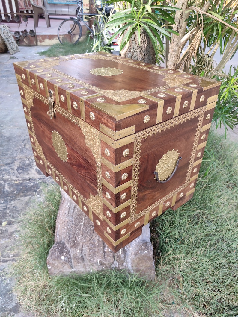 May include: A wooden chest with intricate brass accents and a decorative brass handle. The chest is decorated with floral patterns and has a dark brown finish.
