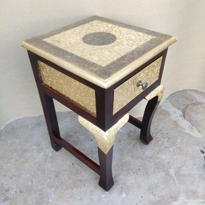 May include: A small wooden side table with a single drawer. The top of the table is decorated with gold and silver inlay. The legs are curved and have gold accents.