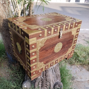May include: A wooden chest with brass accents and intricate carvings. The chest has a hinged lid and a brass lock. The chest is sitting on a tree stump.