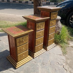 May include: Three ornate wooden pedestals with drawers. The pedestals are painted in shades of red and gold, with intricate floral patterns. Each pedestal has a square top and a base, with multiple drawers. The pedestals are of varying heights.