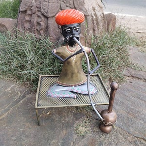 May include: A metal figurine of a person wearing a red turban and a brown robe, sitting on a metal stand and holding a long metal pipe. The figurine is painted with gold and black details.