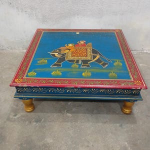 May include: Hand-painted wooden square table featuring an elephant design. The table has a blue top with a red border and gold accents. The elephant is black with gold and red details, and the legs are gold.