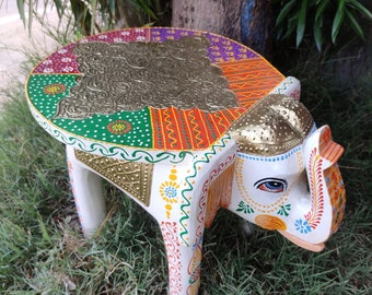 May include: A decorative, hand-painted elephant-shaped side table. The table features a round top with a gold embossed design and colorful, patterned segments. The elephant's body is white with intricate painted details. The table is set on a grassy surface.