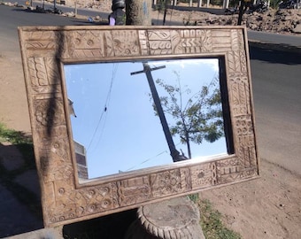 Puede incluir: Un espejo rectangular con marco de madera con intrincadas tallas. El marco es de color marrón claro. El espejo refleja el cielo y un árbol. El espejo está apoyado en un pedestal de piedra al aire libre.