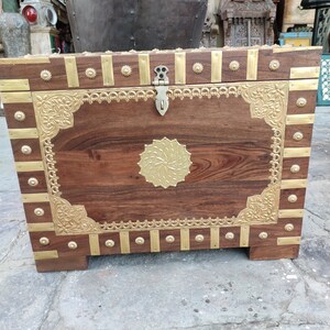 May include: A wooden storage chest with intricate brass accents. The chest has a hinged lid with a brass latch and decorative brass trim around the edges. The chest is sitting on a concrete floor.