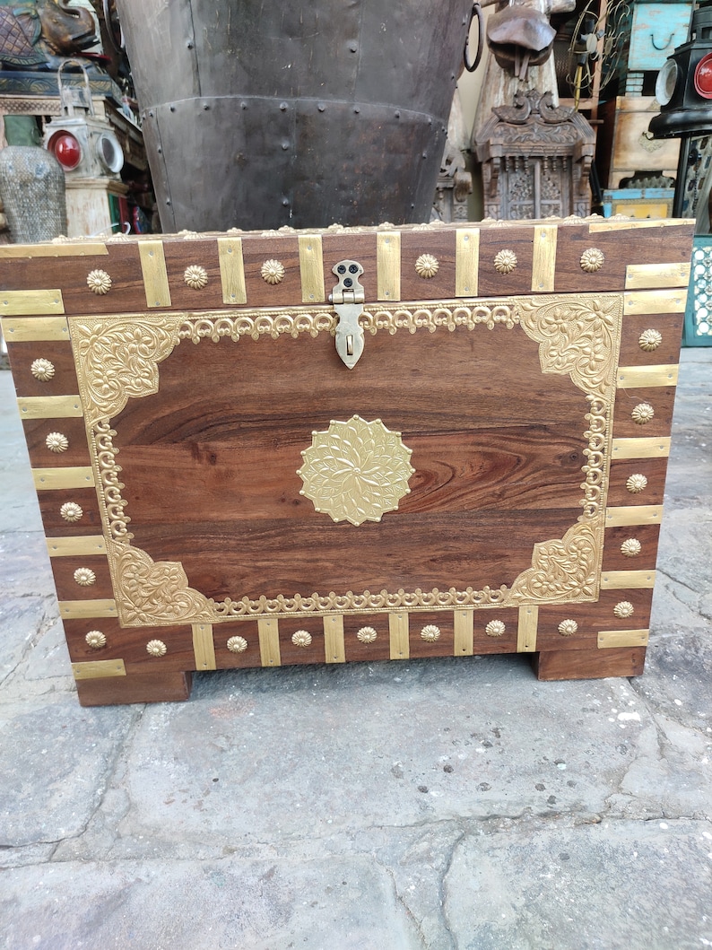 May include: A wooden storage chest with intricate brass accents. The chest has a hinged lid with a brass latch and decorative brass trim around the edges. The chest is sitting on a concrete floor.