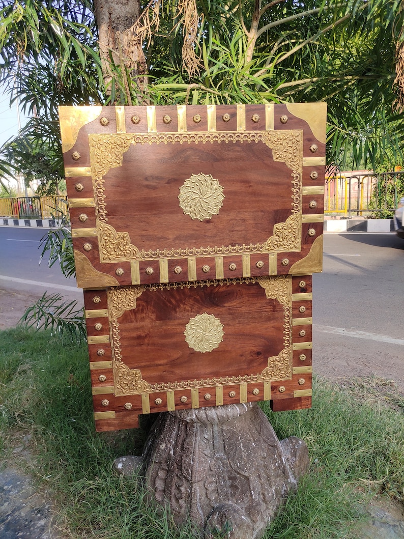 May include: Two wooden boxes with intricate brass inlays. The boxes are stacked on top of each other and have a dark brown finish. The brass inlays feature a floral design and a circular motif in the center.