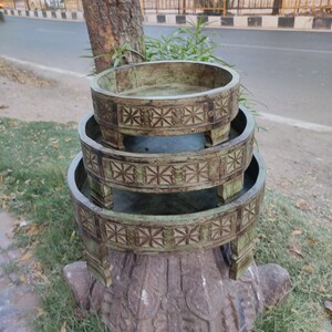 May include: Three-tiered, round wooden serving trays with a distressed green finish. Each tray has decorative carved details around the rim and is supported by short legs. The trays are stacked on a carved stone base.