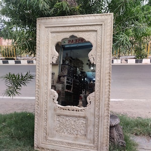 May include: A large, intricately carved wooden mirror with a whitewashed finish. The mirror has a traditional Indian design with a central arch and floral motifs. The mirror is leaning against a tree trunk.