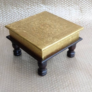 Puede incluir: Una caja de madera cuadrada con tapa dorada y diseños grabados intrincados. La caja se asienta sobre cuatro patas de madera torneadas.