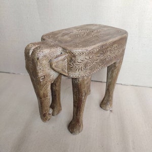 May include: A wooden elephant-shaped stool with carved details and a rectangular top. The stool is painted in a light brown color.