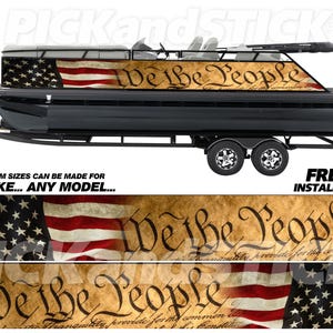 May include: A black pontoon boat with a custom vinyl wrap featuring a faded American flag and the text "We the People" in a vintage style font. The boat is on a trailer with two wheels.