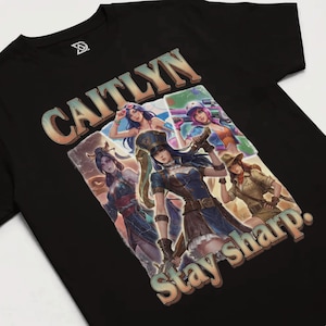 May include: Black t-shirt featuring a colorful graphic of the character Caitlyn from a video game. The graphic includes multiple images of Caitlyn in different outfits, with the text "CAITLYN" and "Stay sharp."