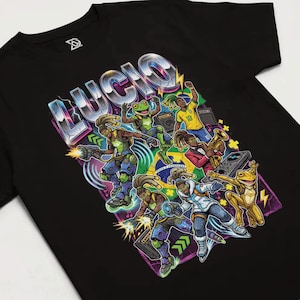 May include: Black t-shirt featuring a vibrant graphic design with the word "LUCIO" in a metallic, stylized font. The design includes multiple cartoon characters, some resembling frogs, and elements of the Brazilian flag.
